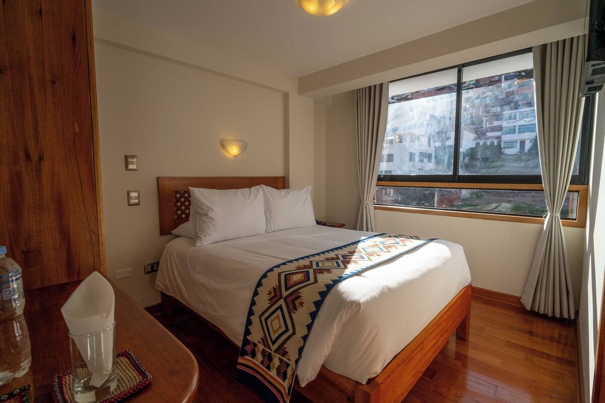 Une chambre d’hôtel avec un lit en bois, une table de chevet avec un verre, et une vue sur les bâtiments par la fenêtre.