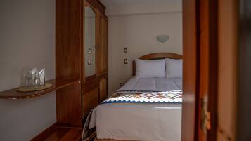 Une chambre à coucher avec une armoire en bois, une table de chevet sur laquelle reposent des lunettes, et un lit recouvert d’une couvre-lit à motifs.