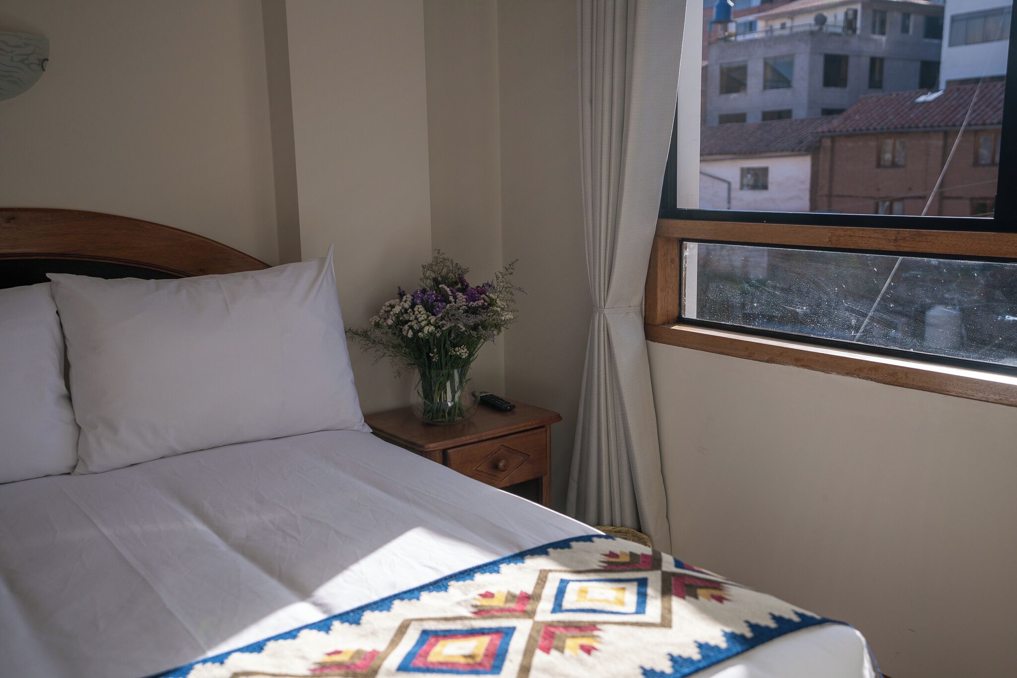 Una cama con sábanas blancas, cabecera de madera, mesita de noche con un jarrón de flores y una ventana con vista a edificios.