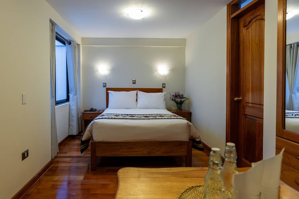 Une chambre d’hôtel avec un lit en bois, des lampes de chevet, une table de nuit, une fenêtre avec des rideaux et une porte en bois.