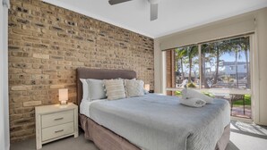 A bedroom with a brick wall, a bed with a headboard, a nightstand with a lamp, and a window with a view of palm trees and a building. - Lakeside Apartments - Ground floor (Merimbula)