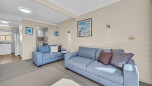 A living room with a blue sofa, a small round table, and a kitchen area visible through an open door. - Lakeside Apartments - Ground floor (Merimbula)