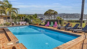 A rectangular swimming pool with a tiled edge, surrounded by a brick deck. There are lounge chairs and colorful bean bags along the poolside. - Lakeside Apartments - Ground floor (Merimbula)