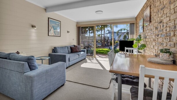 A modern living room with a grey sofa, a wooden dining table, and a stone wall. - Lakeside Apartments - Ground floor (Merimbula)
