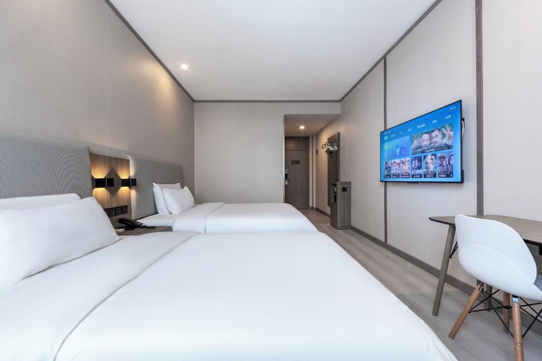 A hotel room with two beds, a desk, a chair, a TV mounted on the wall, and a trash bin.