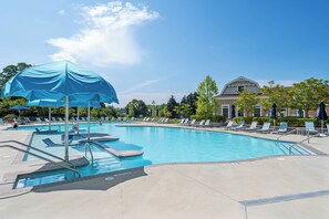 Une piscine entourée d’arbres, avec des chaises longues et des parasols, et un bâtiment visible au loin.
