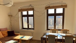 A cozy living room with a sofa, a dining table with chairs, a radiator, and two windows with views of buildings. - Cozy family apartment in the heart of Görlitz, also suitable for fitters (Görlitz)