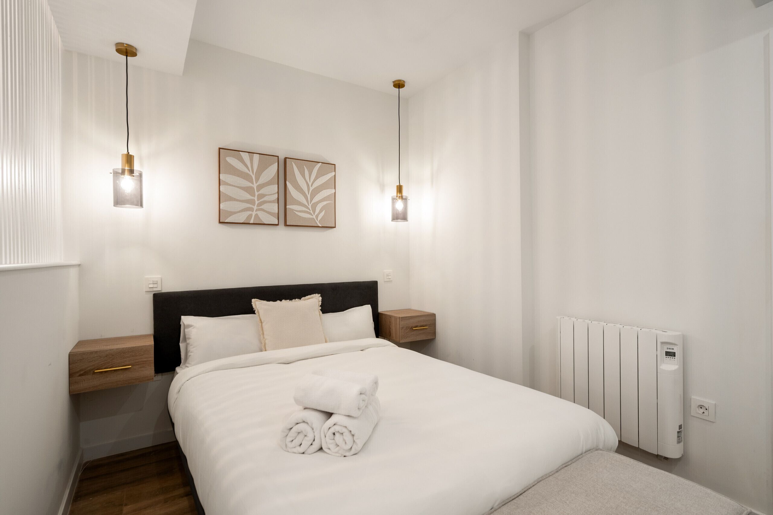 A neatly made bed with white linens and rolled towels, a black headboard, bedside tables with wooden drawers, and two wall-mounted light fixtures.