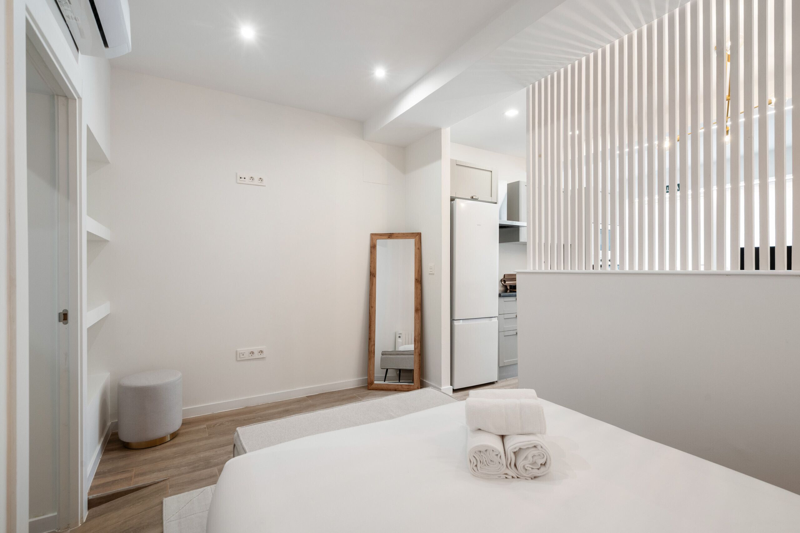 A modern hotel room with a large bed, a small round stool, a mirror, and a kitchenette in the background.