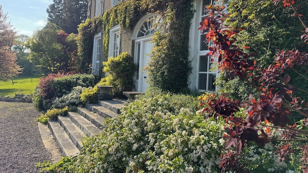 En bygning med en stenfassade, store vinduer og en velholdt have med forskellige planter samt en grussti, der fører til indgangen.