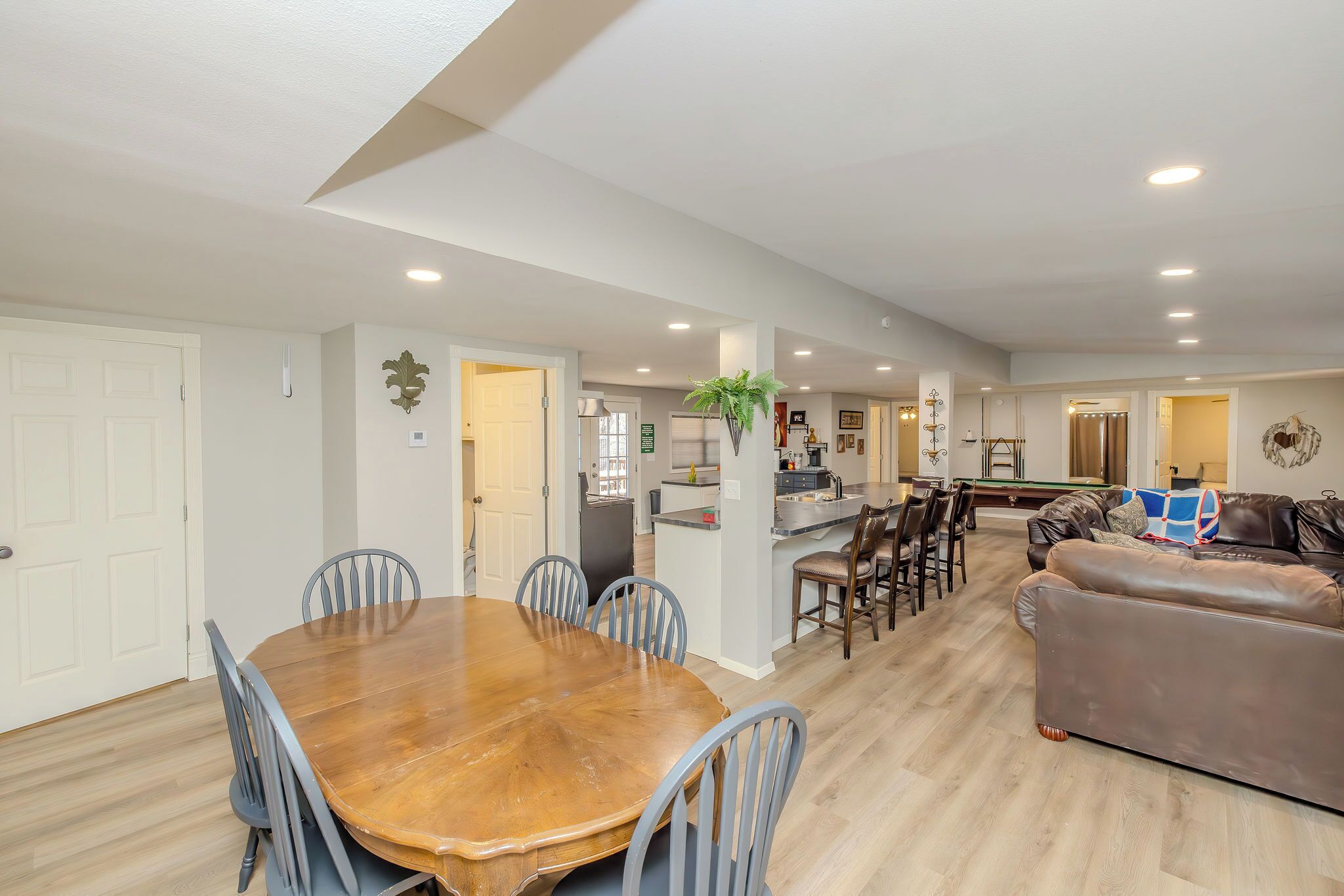 A spacious living area with a wooden dining table, blue chairs, a brown leather sofa, and a kitchen in the background.