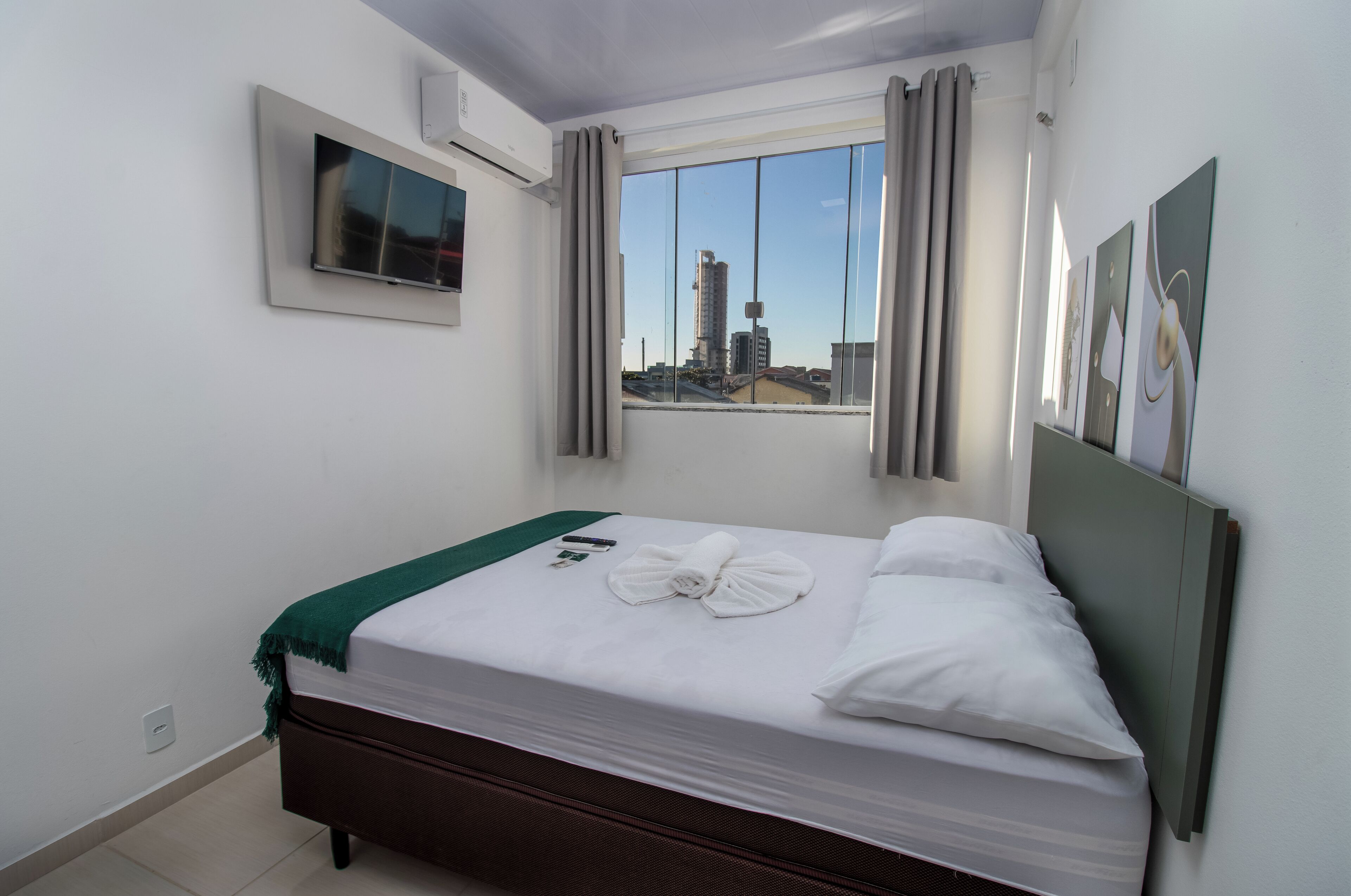 A neatly made bed with a green throw, a flat-screen TV mounted on the wall, and a window with curtains.
