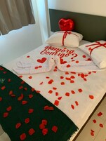 Una cama con sábana blanca, almohadas con forma de corazón rojo y un mensaje en rojo.