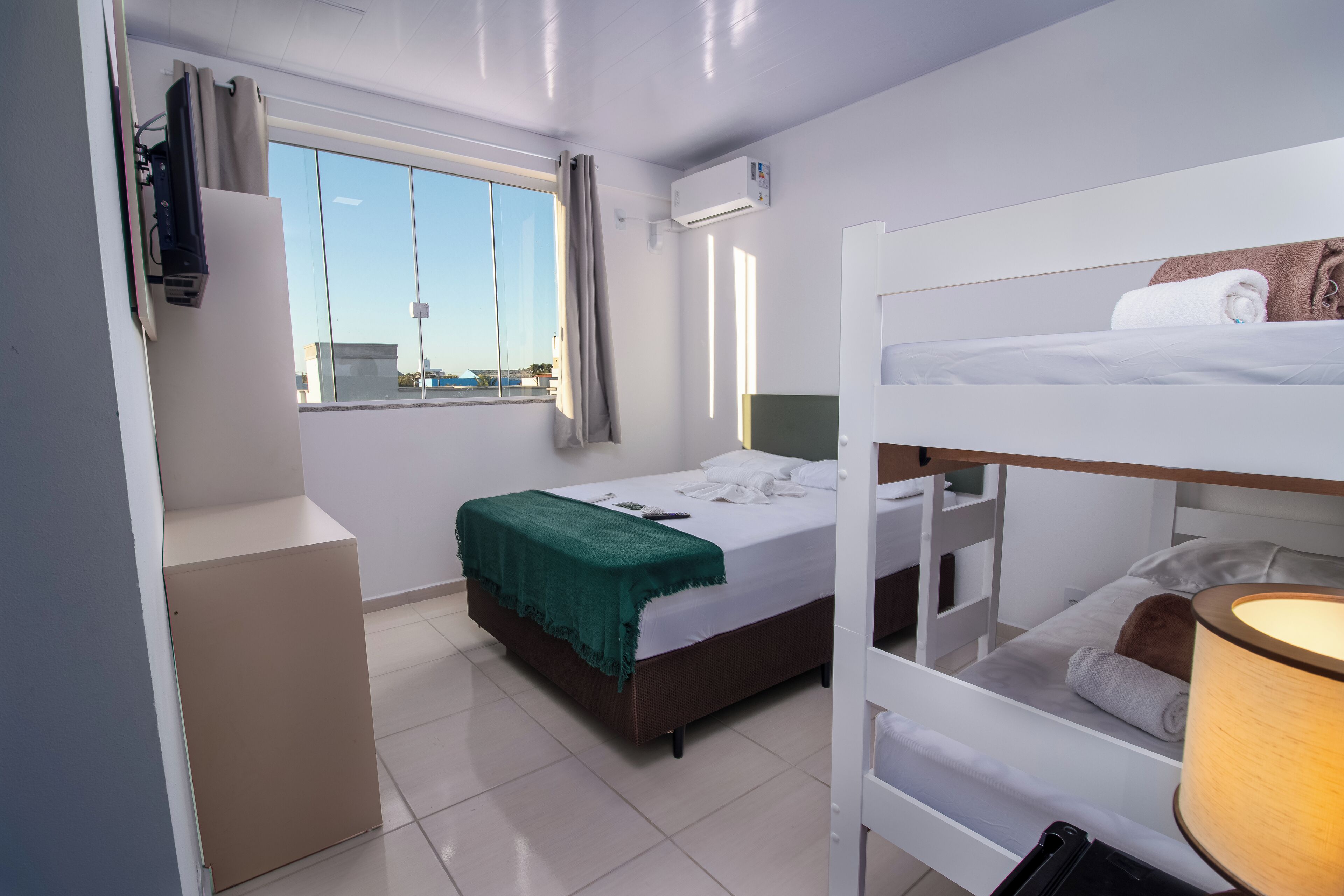 Habitación de hotel compacta con literas, una cama individual con una manta verde, una ventana con cortinas y vistas a la ciudad.