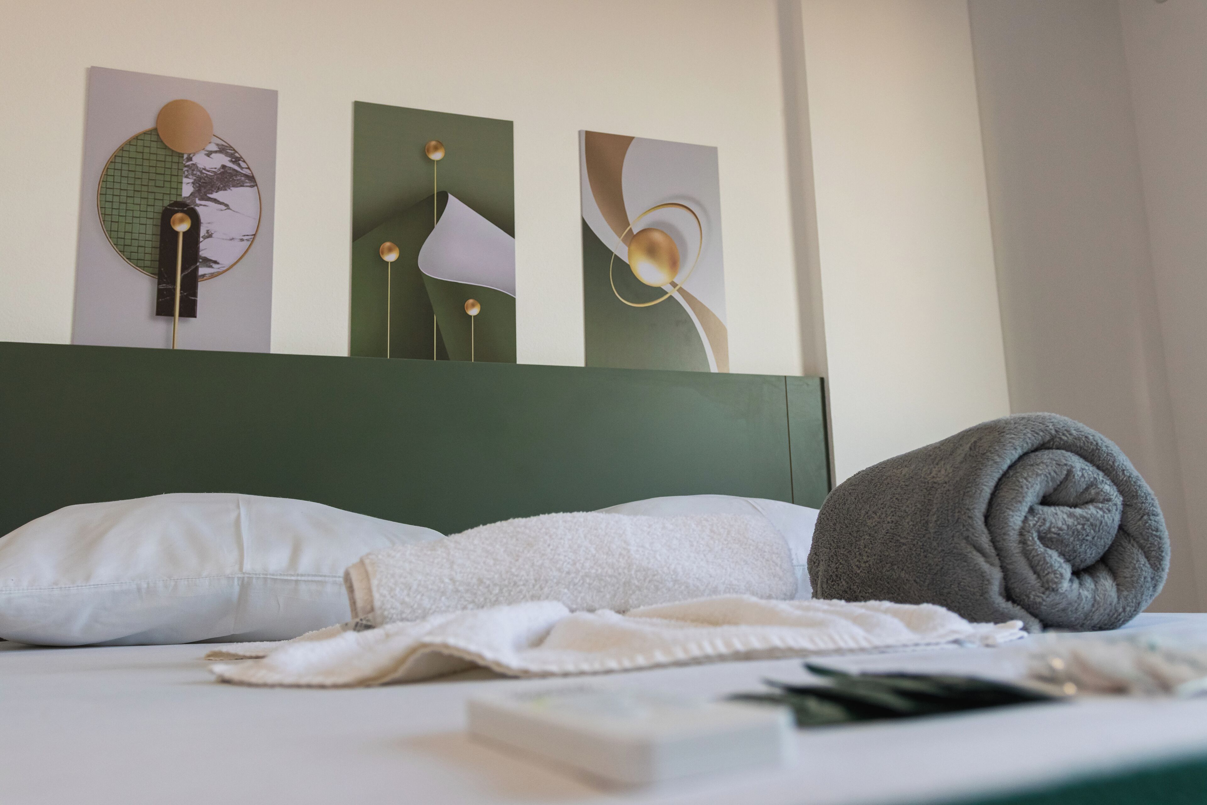 A neatly made bed with a white pillow, a rolled gray towel, and a white towel on a white bedspread. There are abstract wall art pieces above the bed.