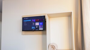 A wall-mounted TV displaying a streaming service interface with Netflix and Disney+ icons, a white wall, and a shelf with a rolled towel. - Pousada Paraná (Penha)