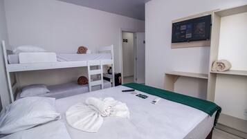 A hotel room with a bunk bed, a single bed, a TV mounted on the wall, and a small shelf with towels.