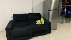 A black sofa with a plush toy and a stainless steel refrigerator in a room with white walls and a tiled floor. - Pousada Paraná (Penha)