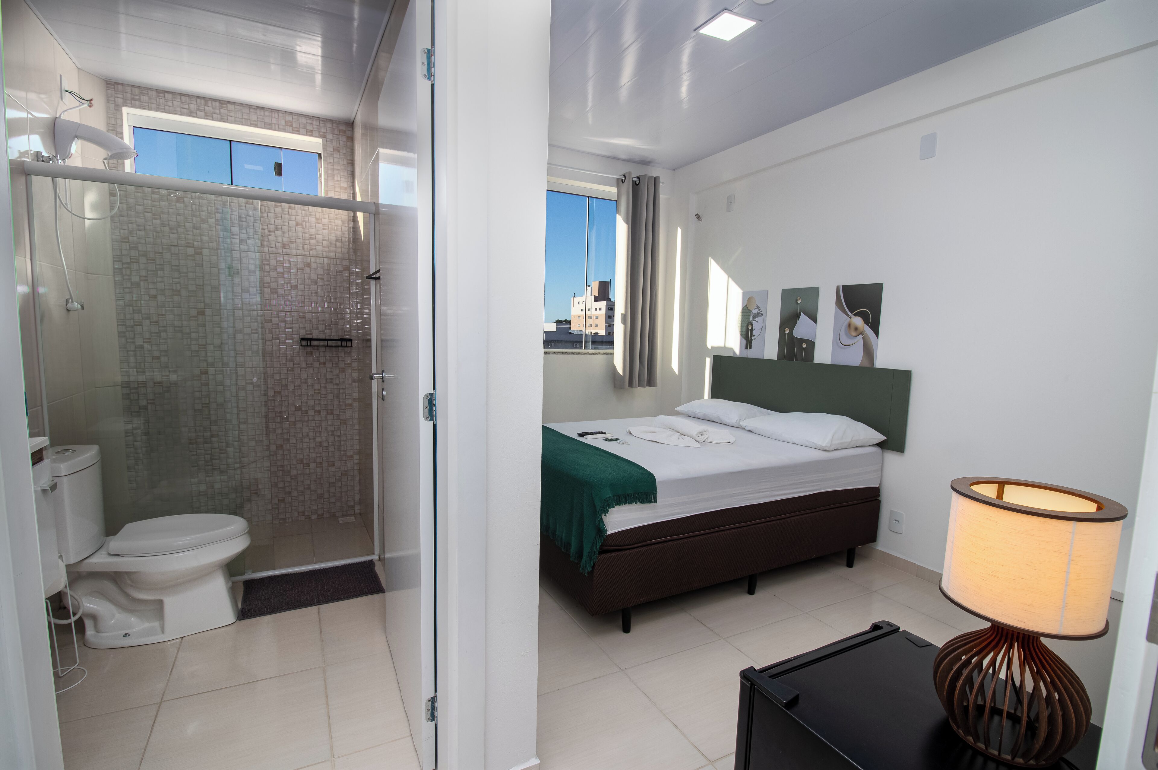 A hotel room with a glass-enclosed shower, a bed with a green throw, a bedside lamp, and a view of the city.