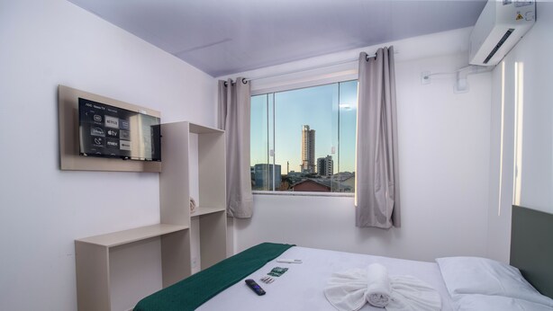 A hotel room with a bed, a TV mounted on the wall, a window with curtains, and a city view. - Pousada Paraná (Penha)