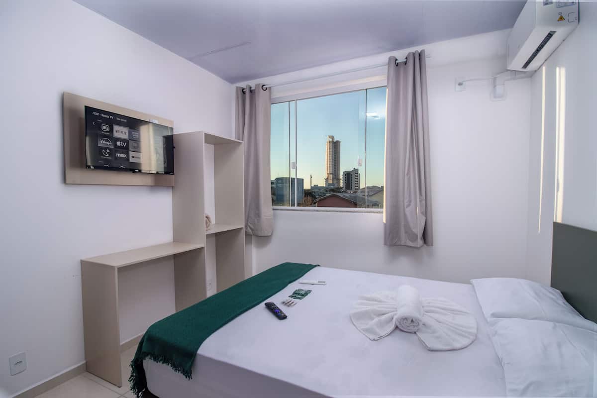 A hotel room with a bed, a TV mounted on the wall, a window with curtains, and a city view.