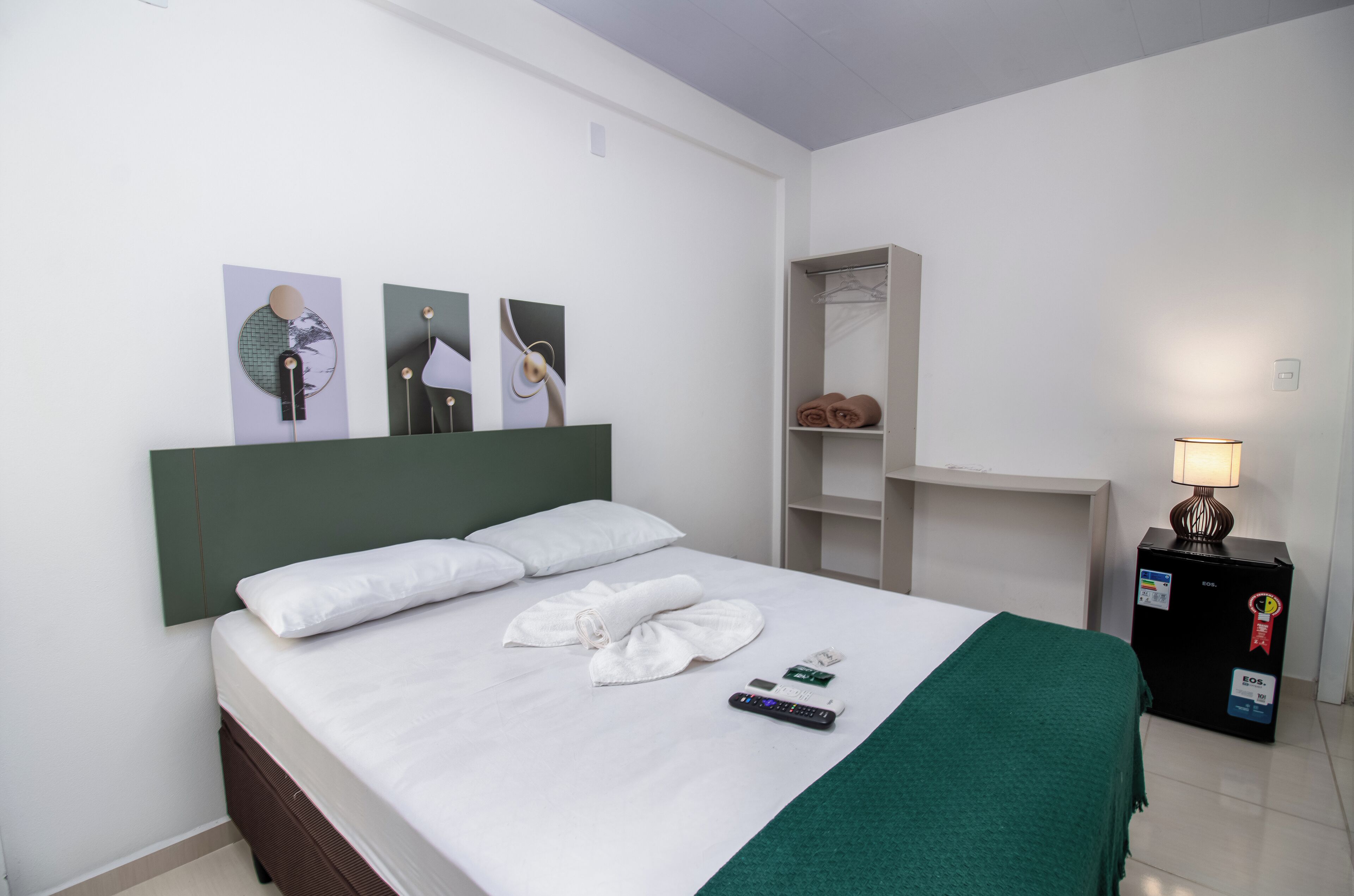 A neatly made bed with white linens, a green throw blanket, and a remote control on the bed. There is a wall-mounted shelf with folded towels and a lamp on a table beside the bed.