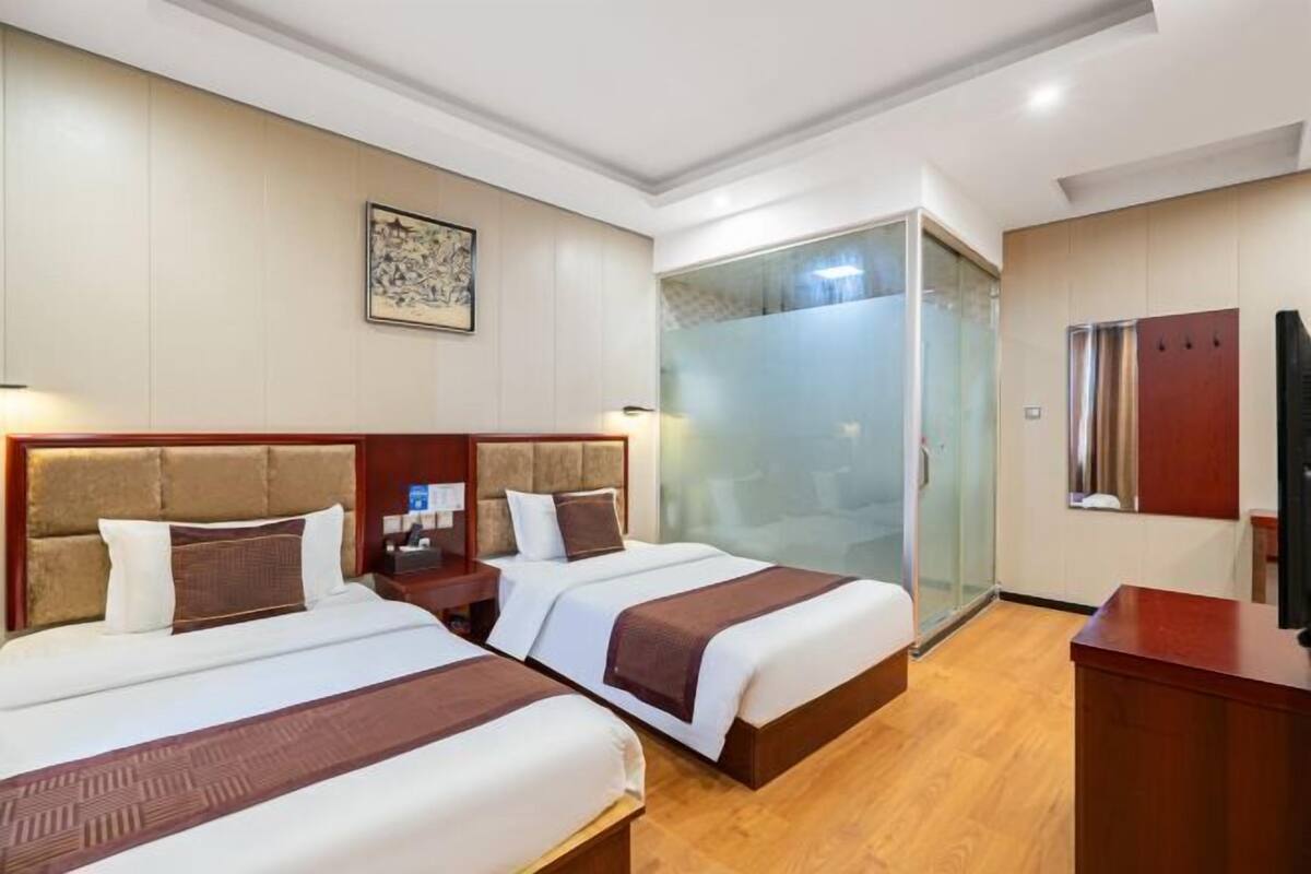 A hotel room with two beds, a wooden headboard, a glass shower enclosure, and a wooden desk.