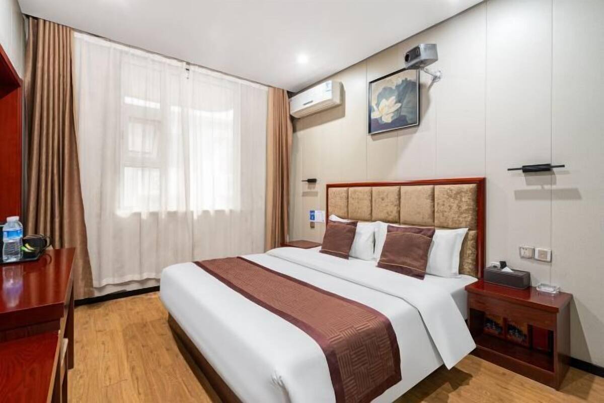 A hotel room with a large bed, a wooden nightstand, a desk with a chair, a window with curtains, and an air conditioning unit.