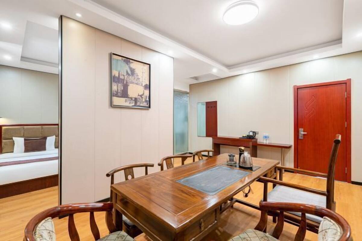 A hotel room with a wooden dining table, chairs, a bed, a desk with a chair, and a red door.