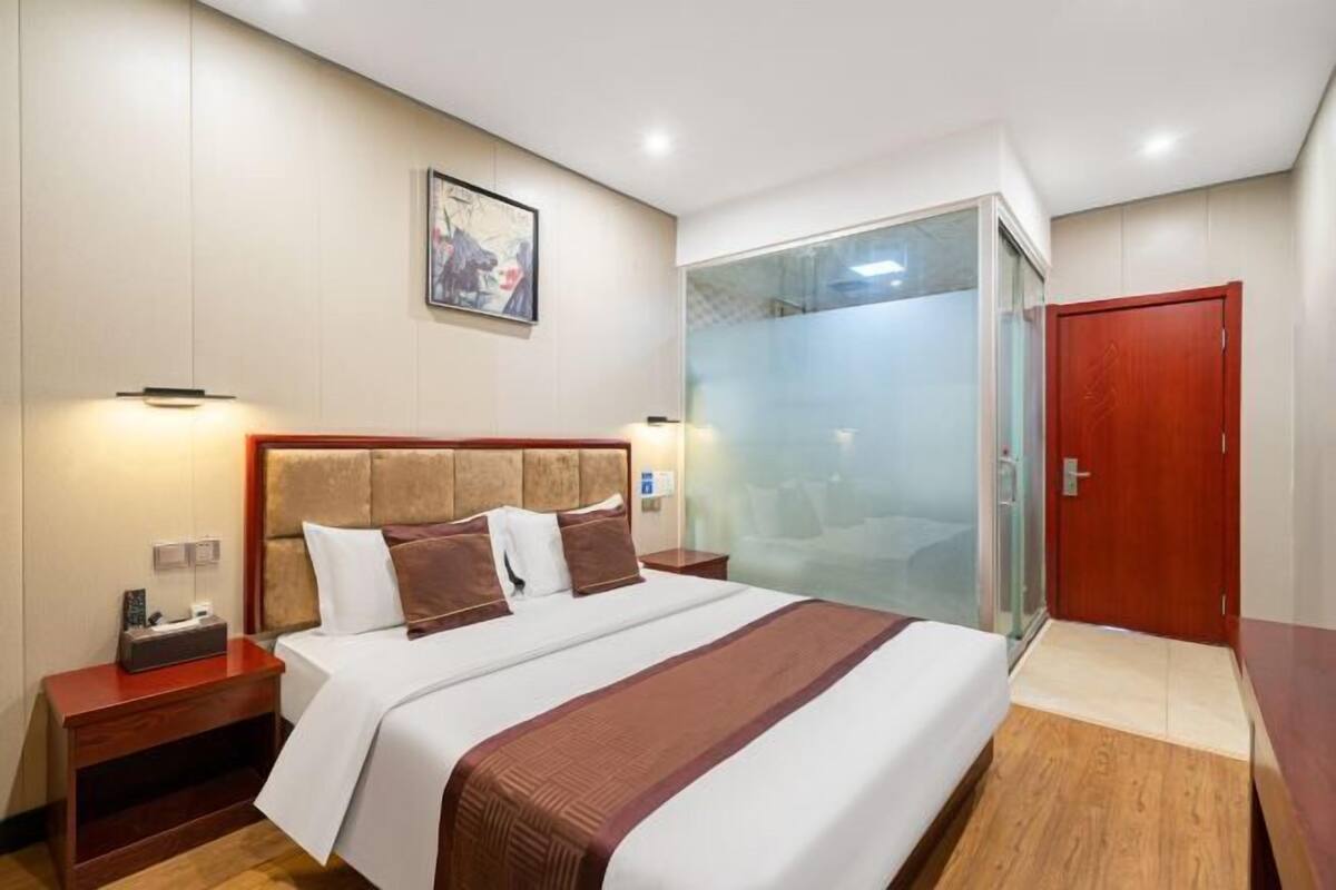 A hotel room with a bed, bedside tables, a nightstand, a glass partition, and a wooden door.