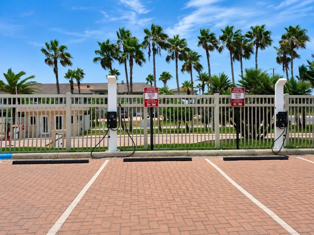A parking area with electric vehicle charging stations, a white fence, and palm trees in the background.