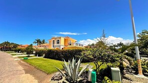 A large, yellow building with a balcony, surrounded by well-maintained gardens and a clear sky.