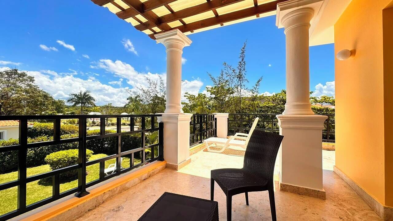 Un balcón con columnas blancas, una silla negra y vistas del entorno cubierto de vegetación.