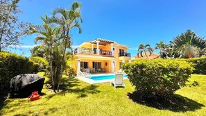 A two-story house with a swimming pool, surrounded by lush greenery and palm trees.