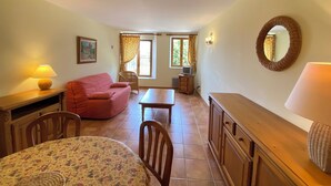 Un salon confortable avec un canapé, une table à manger et un téléviseur. Il y a des fenêtres avec des rideaux, une lampe et un miroir au mur.