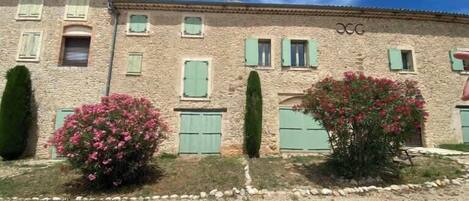 A stone building with multiple windows and shutters, a gravel driveway, and a well-maintained garden with pink flowers.