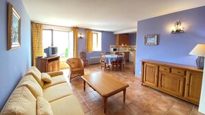 A living room with a sofa, a wooden coffee table, and a dining area with a table and chairs. There is a kitchen area with cabinets and a microwave.