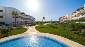 A swimming pool surrounded by a well-maintained lawn, with a clear sky and a row of white buildings in the background. - Apartamento Vamos (Mar de Pulpi) (San Juan de los terreros)