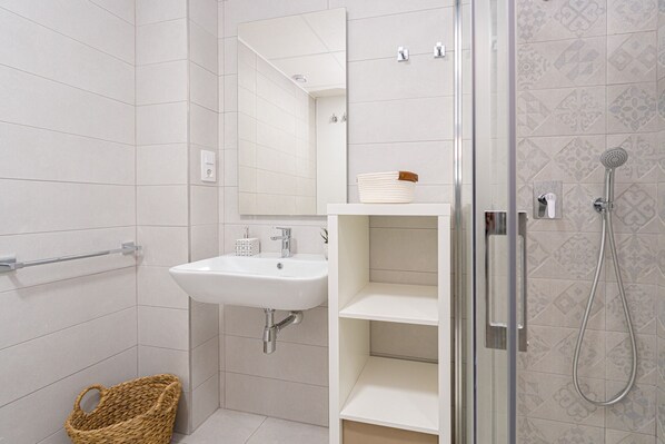 A bathroom with a white sink, mirror, and shower. There is a basket on the floor and a shelf with a white container. - Apartamento Vamos (Mar de Pulpi) (San Juan de los terreros)