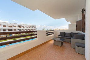Un balcón con sofá, una mesita y vistas a una piscina y edificios.