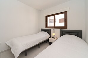 Una habitación pequeña y limpia con dos camas individuales, una mesita de noche con lámpara y una ventana con persianas.