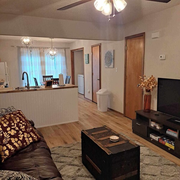 Sala de estar con un sofá de cuero marrón, una mesa de centro de madera, un televisor sobre un mueble bajo, una zona de cocina con una mesa de comedor y sillas, y un ventilador de techo.