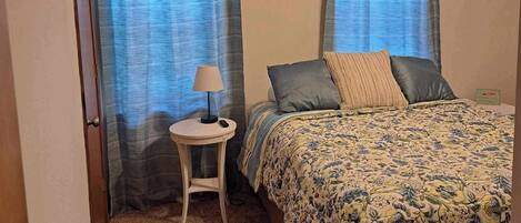 Ein Schlafzimmer mit einem Bett, einem kleinen Nachttisch mit Lampe und zwei mit Vorhängen versehenen Fenstern.