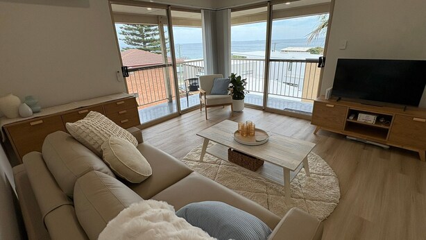 Un salón moderno con sofá, mesa de centro y televisor, con vistas a un balcón y al océano.