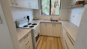 Una cocina compacta con armarios blancos, estufa, lavamanos y ventana con vistas a palmeras.
