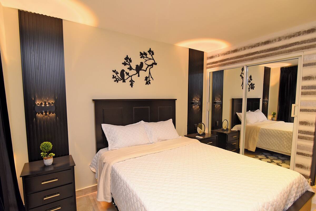 A hotel room with a large bed, a nightstand, a dresser, a mirror, and a wall with a decorative tree design.