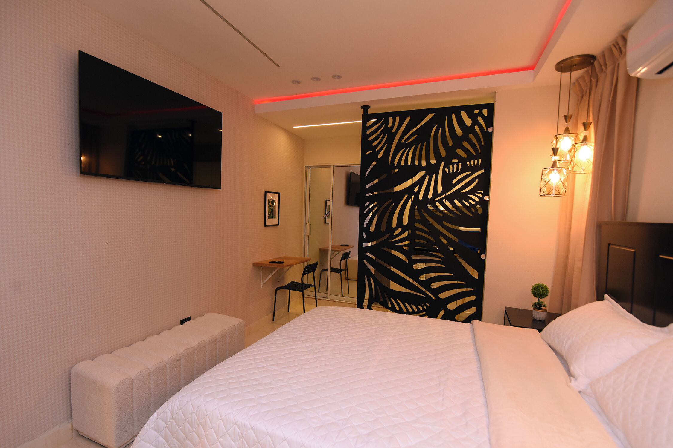 A modern hotel room with a large bed, a flat-screen TV mounted on the wall, a small desk with a chair, and a decorative partition with leaf patterns.