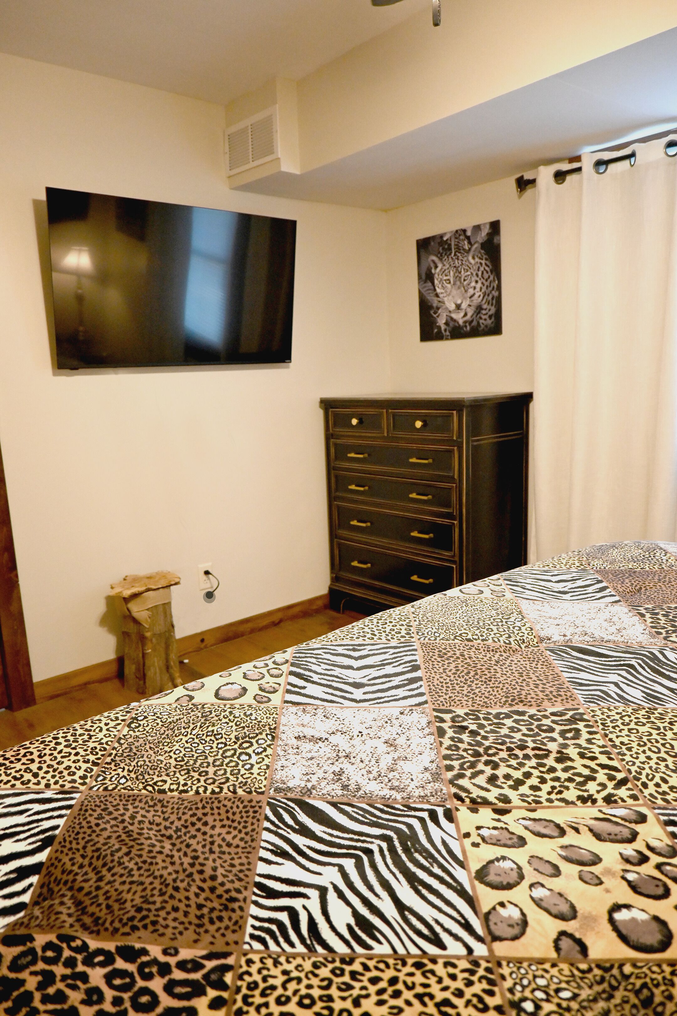 Una habitación con colcha de estampado de cebra en la cama, cómoda de madera oscura, televisor montado en la pared e imagen con estampado de leopardo.