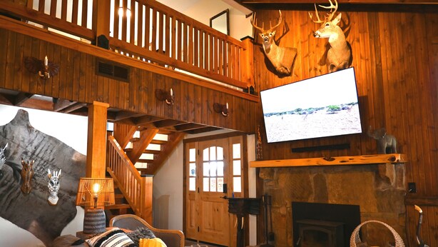 A cozy cabin interior with a fireplace, wooden walls, a mounted deer head, and a television.