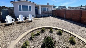 A residential property with a gravel driveway, a small shed, and a wooden fence. There are three white Adirondack chairs and a small garden area with shrubs and flowers.